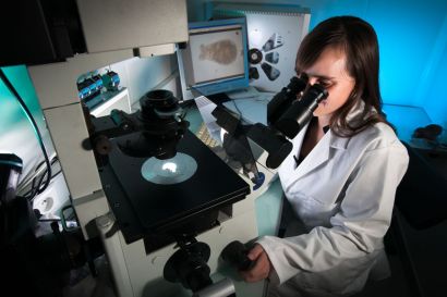Une jeune femme en blouse regarde dans un microscope. On voit sur un écran les champignons qu'elle observe.