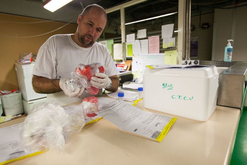 Un homme portant des gants en train de réceptionner des boites contenant des prélèvements biologiques. Il a un stylo dans la main et il y a des feuilles sur le plan de travail devant lui.