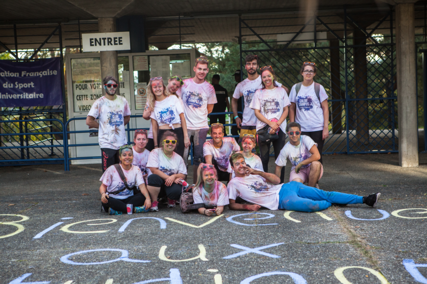 Un groupe d'étudiants couverts de poudre colorée qui posent devant un marquage à la craie au sol indiquant "bienvenue aux étudiants"