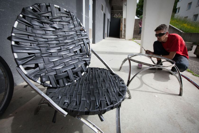 Au premier plan : un fauteuil à partir de roues et chambres à air. Au second plan un jeune homme bricole.