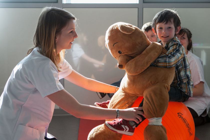 Un petit garçon tient un énorme nounours sur un gros ballon. Une étudiante en blouse souriante lui tient les jambes.