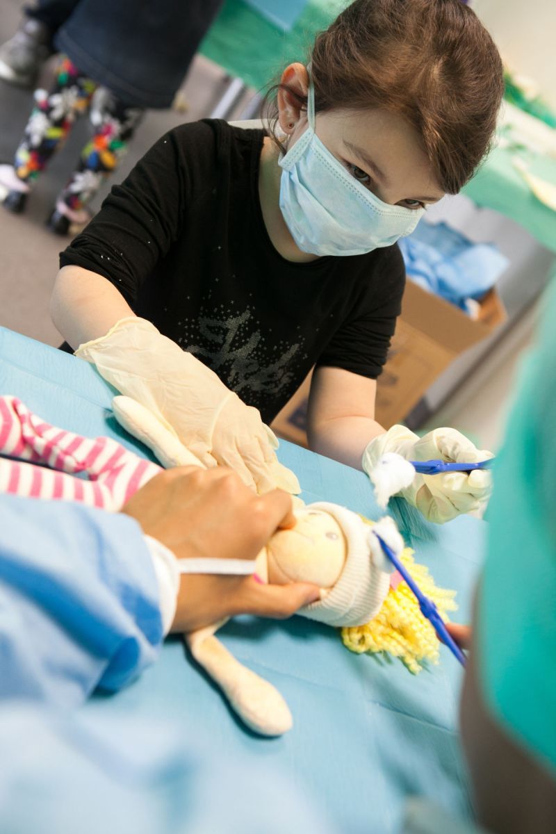 Bloc opératoire : une petite fille équipée d'un masque et de gans en latex tient une pince avec du coton pour désinfecter sa poupée.