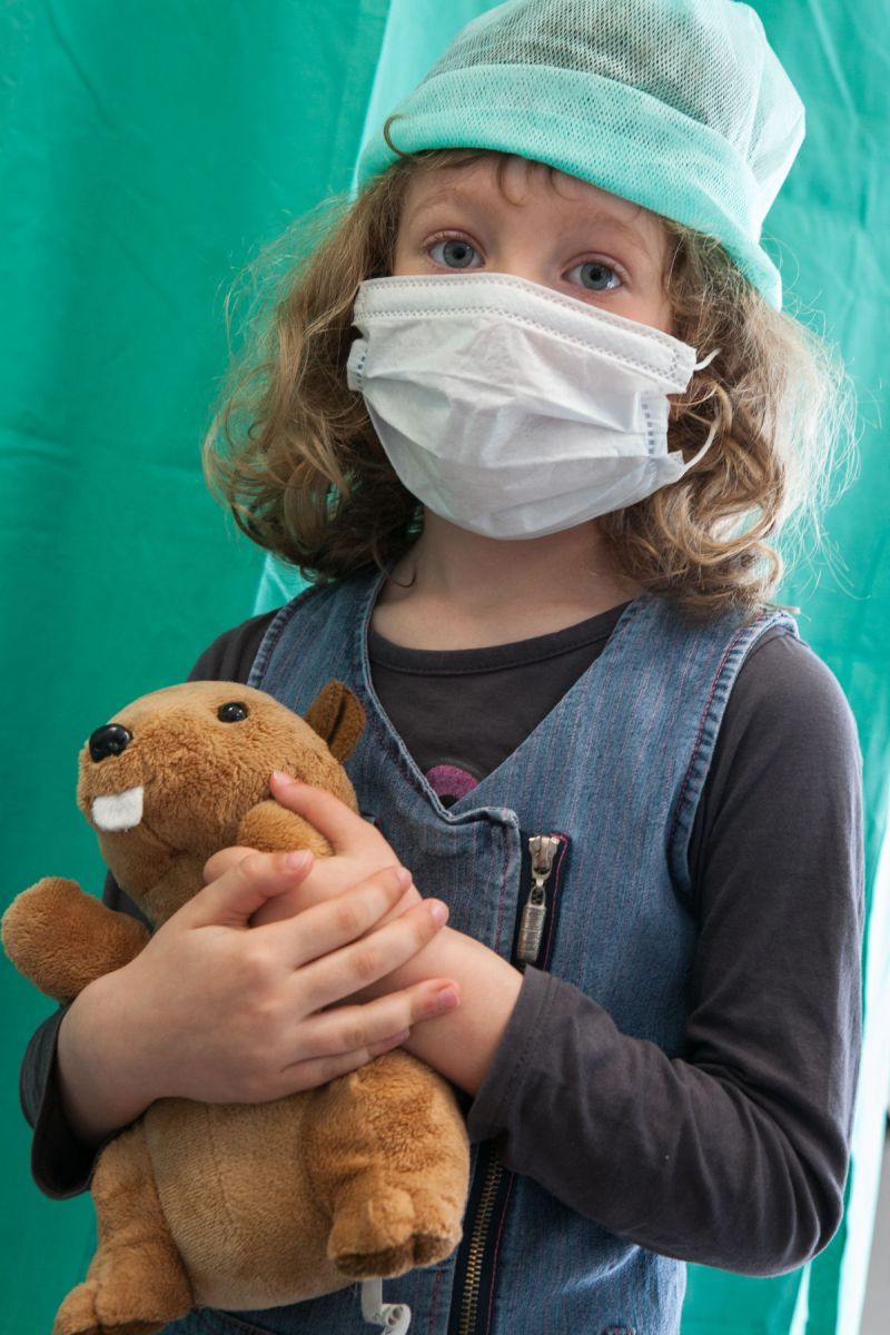 Une petite fille tient son nounours. Elle porte un masque et une charlotte avant d'entrer au bloc opératoire.