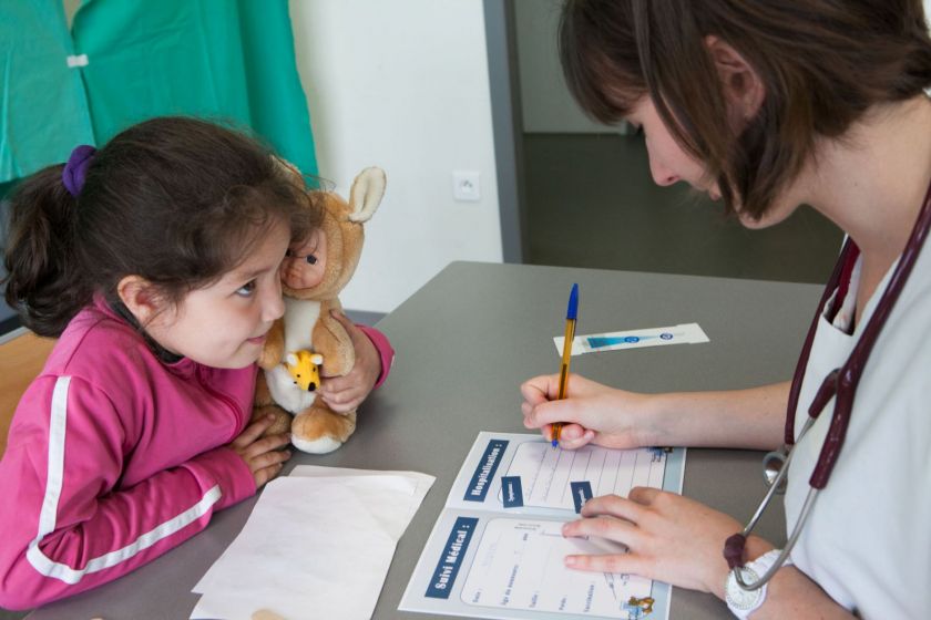 Une petite fille tient son nounorus assise à une table en face d'une jeune femme équipée d'un stétoscope qui remplit un carnet.