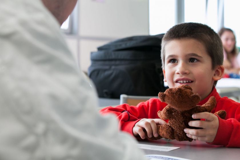 Un petit garçon souriant assis à une table présente son nounours à un adulte de dos.