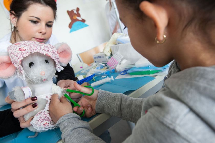 Une petite fille coupe un bandge sur une souris en peluche tenue par une étudiante.