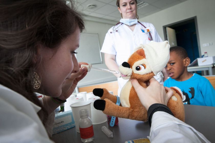 Une jeune fille en blouse donne des gouttes à un nounours bambi sous l'oeil inquiet d'un petit garçon.