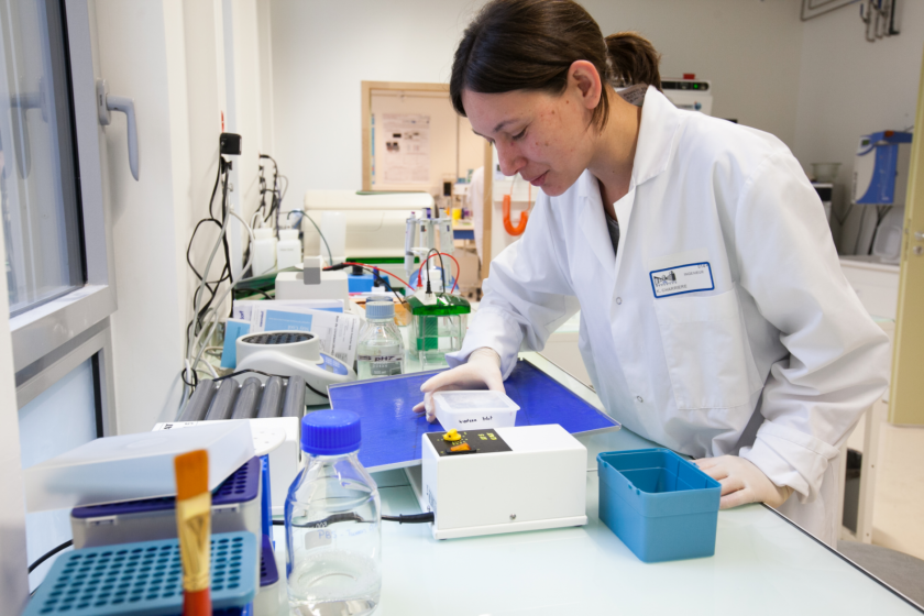 Dans un laboratoire, une jeune femme en blouse en train d'examiner le contenu d'une boite en plastique au dessus d'un appareil évoquant une sorte de balance.