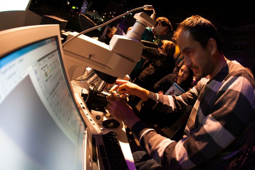 Un homme place un matériau sous un microscope. Au premier plan un écran d'ordinateur et du public en arrière plan. 