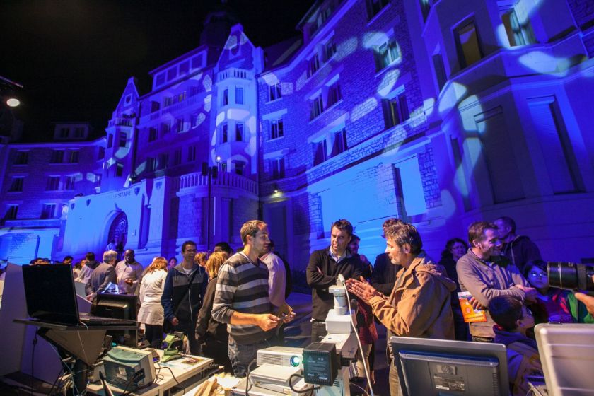 Des gens groupés autour des stands devant le bâtiment Canot éclairé avec projection d'images. 