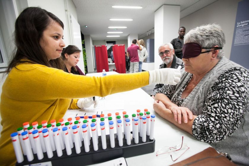 Une étudiante fait renifler un échantillon à une dame aux cheveux blancs les yeux bandés.