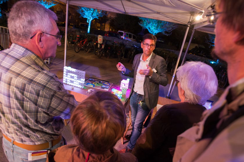 Benoit Piranda devant un stand où sont posées des briques lumineuses parle à du public de dos.