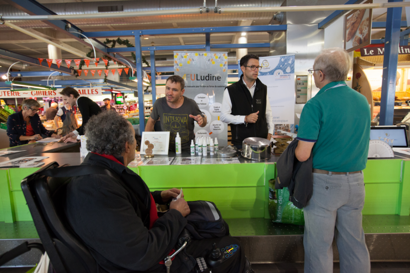 Un stand tenu par des chercheurs au marché avec deux visiteurs devant dont un en fauteuil roulant.