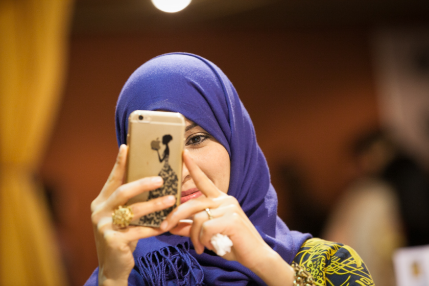 Une femme portant un foulard violet en train de prendre une photo avec son téléphone portable en souriant.