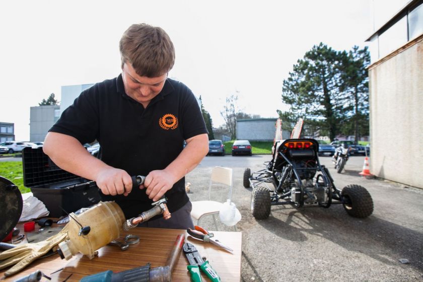 Un éudiant en train de faire de la mécanique à l'extérieur de l'IUT. Un kart en arrière plan.