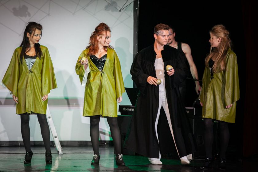 Scène de théâtre : un homme en manteau noir avec une couronne donne des pommes à trois jeunes femmes en blouse verte.