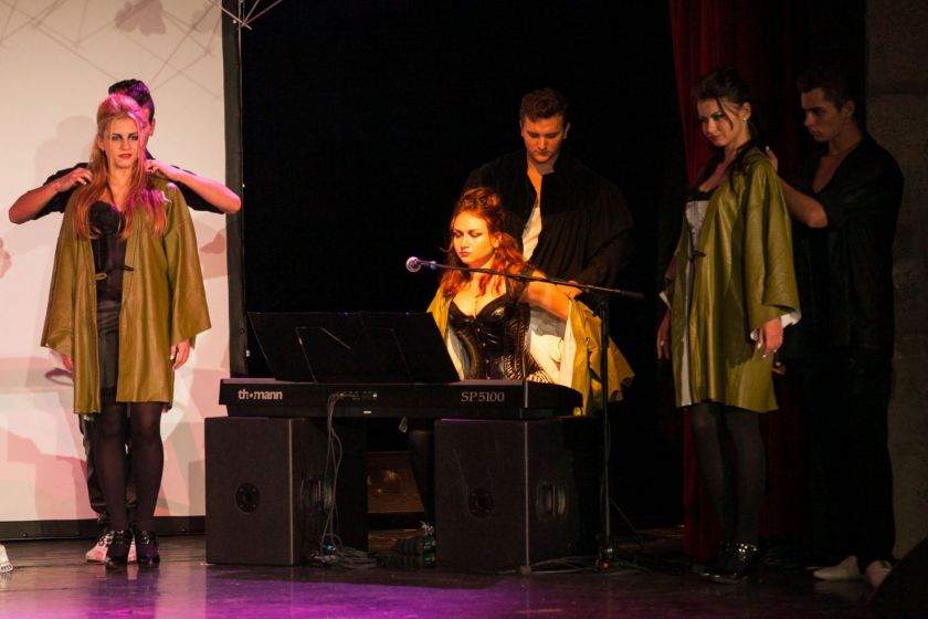 Scène de théâtre : trois jeunes hommes habillent trois jeunes femmes d'une blouse verte. L'une est installée à un clavier.