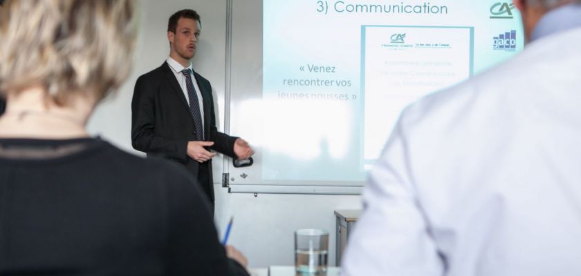 Un étudiant en costume cravate fait une présentation. Deux membres du jury sont de dos.