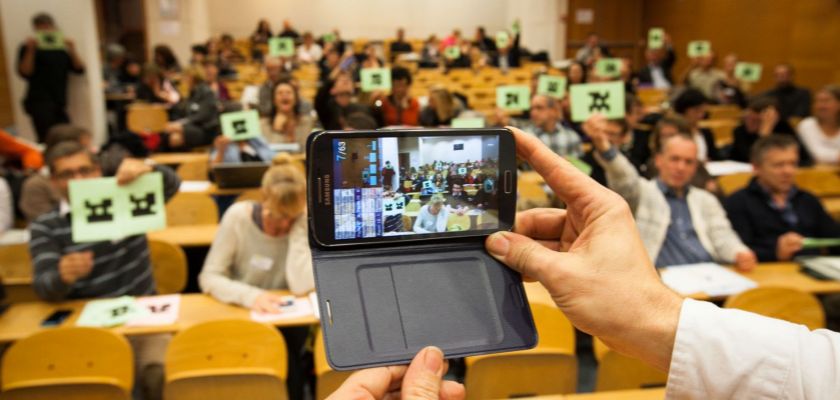 Un amphithéâtre où les gens brandissent des papiers, devant des mains tiennent un téléphone portable.