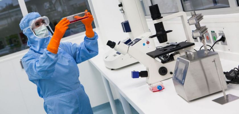 Une femme en combinaison bleue et gants oranges tient une boite avec un liquide rouge, devant une paillasse où se situent sifférents appareils dont un gros microscope.