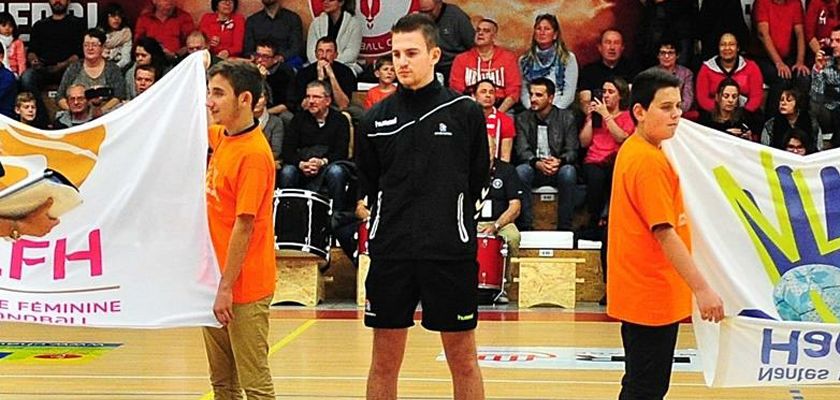 Pierre Vauchez en habit d'arbitre sur un terrain de hand entouré par des jeunes qui portent un drapeau