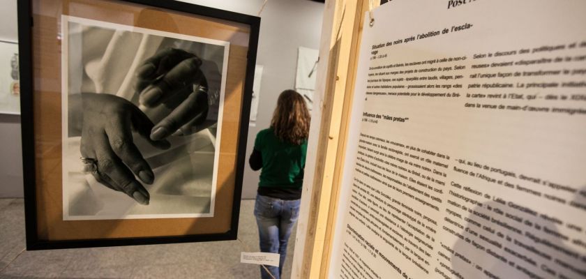 Vue de l'exposition, gros plan sur une photo de mains et un panneau, une femme de dos dans la salle en arrière plan.