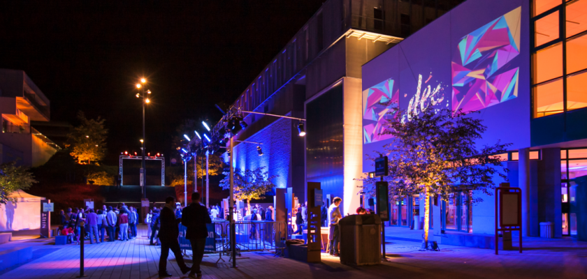 Vue extérieure du parvis de la faculté de médecine de Besançon, de nuit, avec des éclairages bleu violets. Le mot "idées" et des formes géométriques sont projetées sur la façade. Il y a du monde sur la place.