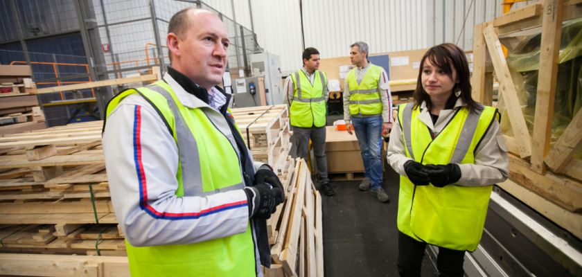 Un homme et une étudiante portant des gilets jaunes fluorescents et des gants dans un atelier. Deux hommes équipés de la même manière discutent à l'arrière plan.