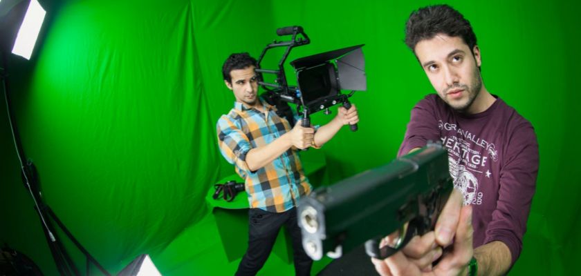 Un jeune homme tient une caméra pointée sur un autre qui tient une arme. En arrière plan, un fond vert et des éclairages de cinéma.