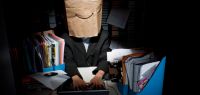 Un homme en costume dans un bureau sombre avec un sace en papier sur la tête et un sourire dessiné sur le sac.